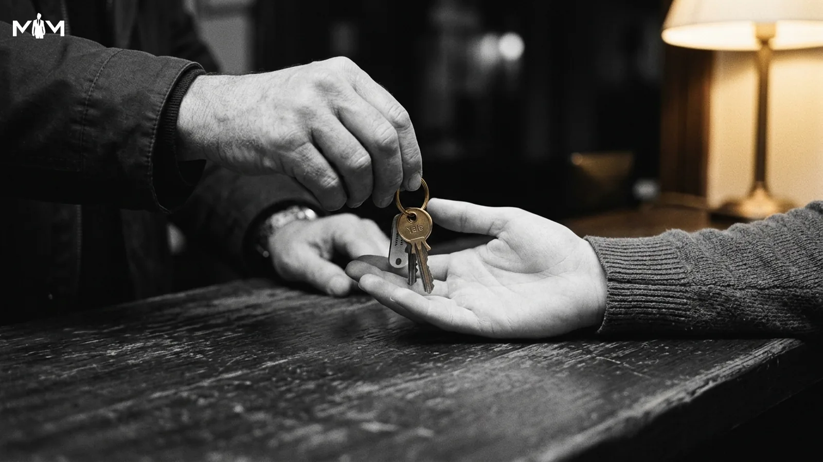 Landlord handing physical keys across a bare countertop to a new tenant warm directional lamplight